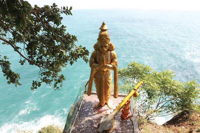 Golden coastal shrine statue overlooking shimmering turquoise ocean waters framed by tropical trees.