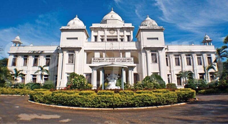 Grand white colonial building with columned entrance, circular driveway, and landscaped flower garden.