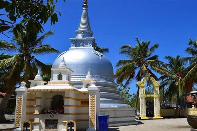 White Buddhist stupa surrounded by coconut palms beneath a clear tropical sky.