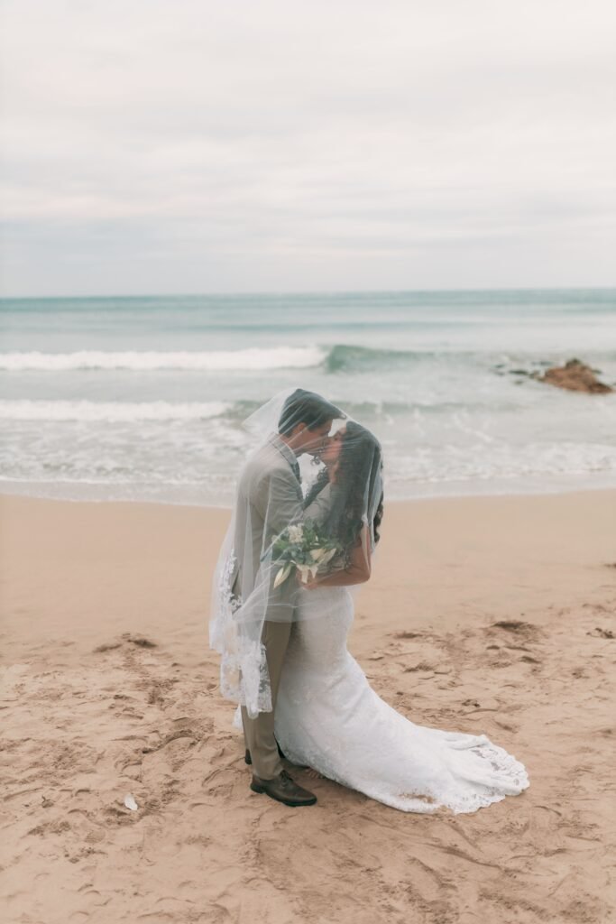 Newlywed couple kisses beneath a flowing veil on a romantic beach by the ocean.