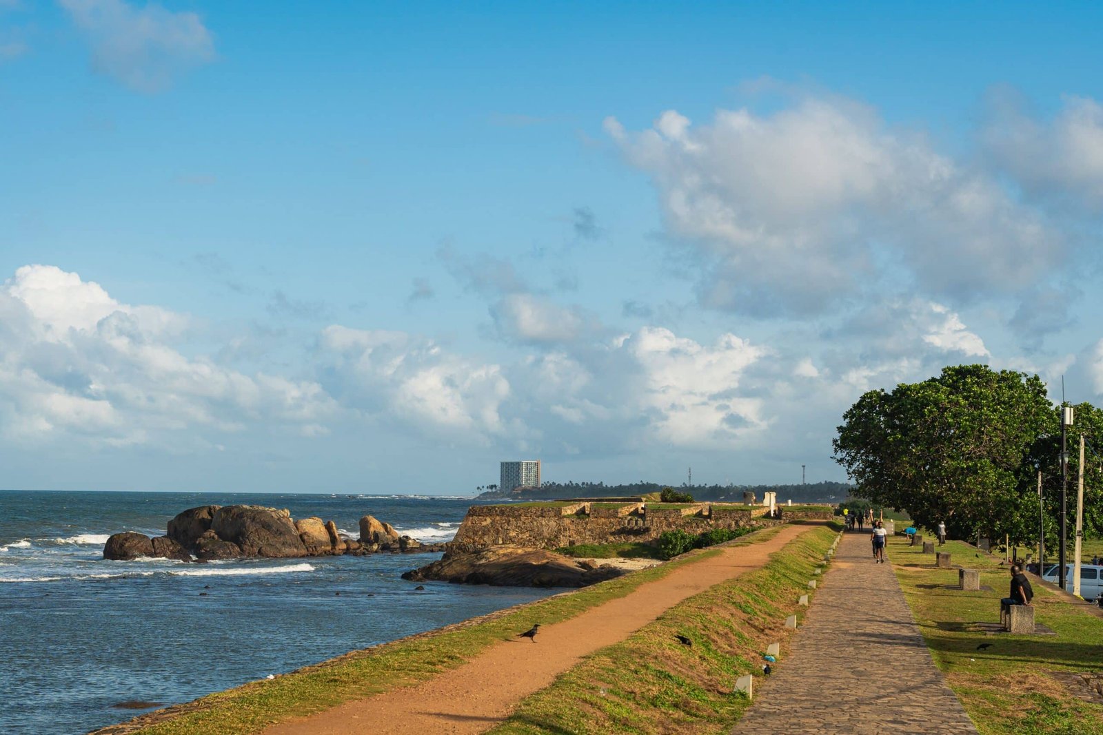 Rocky coastal path beside calm blue water under a bright, cloud-filled sky.