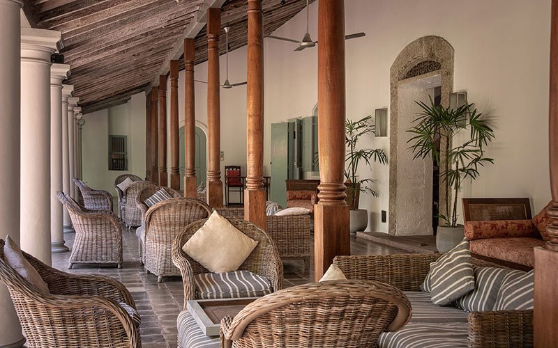 Elegant colonial veranda with wicker seating, wooden columns, green shutters, and tropical plants.