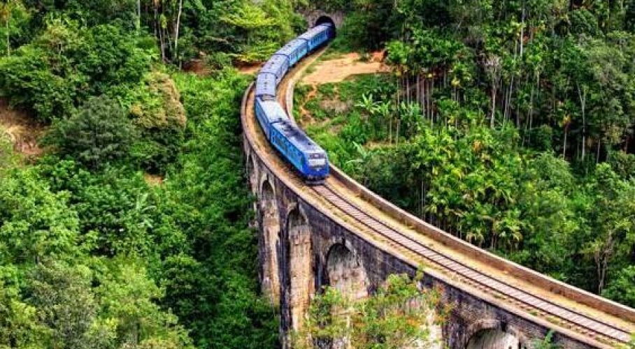 Blue passenger train curves across a historic viaduct surrounded by dense tropical forest.