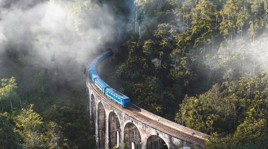 Blue passenger train crosses a misty stone viaduct through lush mountain forest.