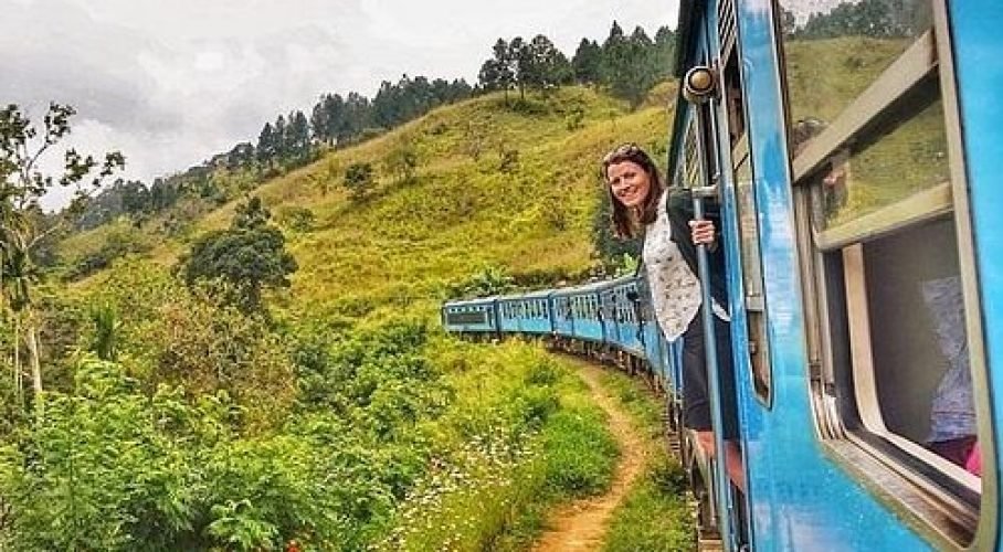 Smiling woman leans from a blue passenger train winding through lush green hills.