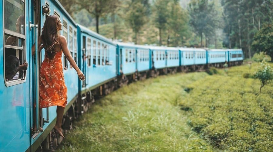 Woman in floral dress rides blue train through lush tea fields.