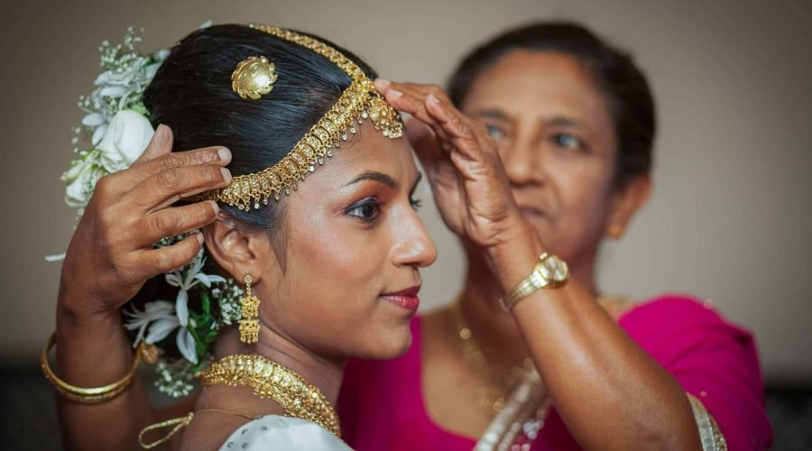 Woman adjusts traditional gold jewelry on bride during wedding preparation.