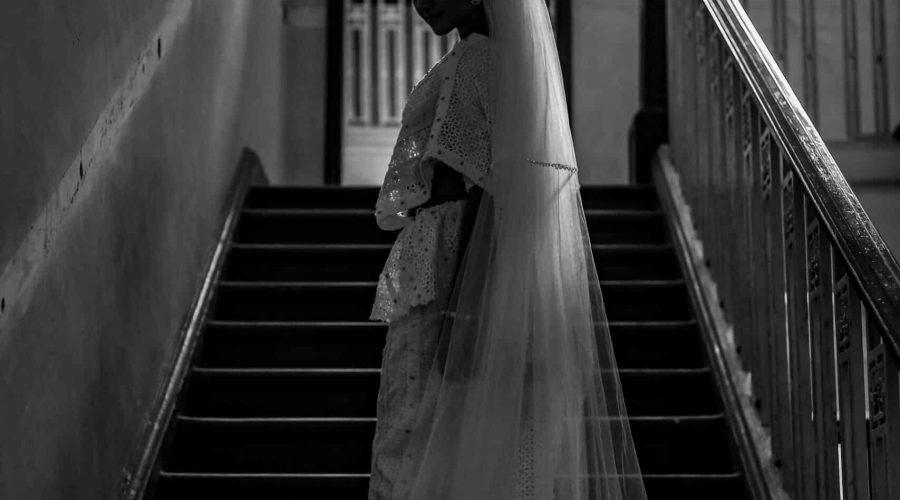 Vintage bride in a flowing gown and veil standing on a shadowy staircase.