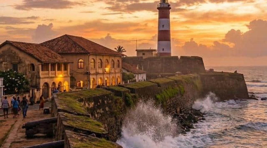 Historic coastal lighthouse and stone fort overlooking crashing waves at sunset.