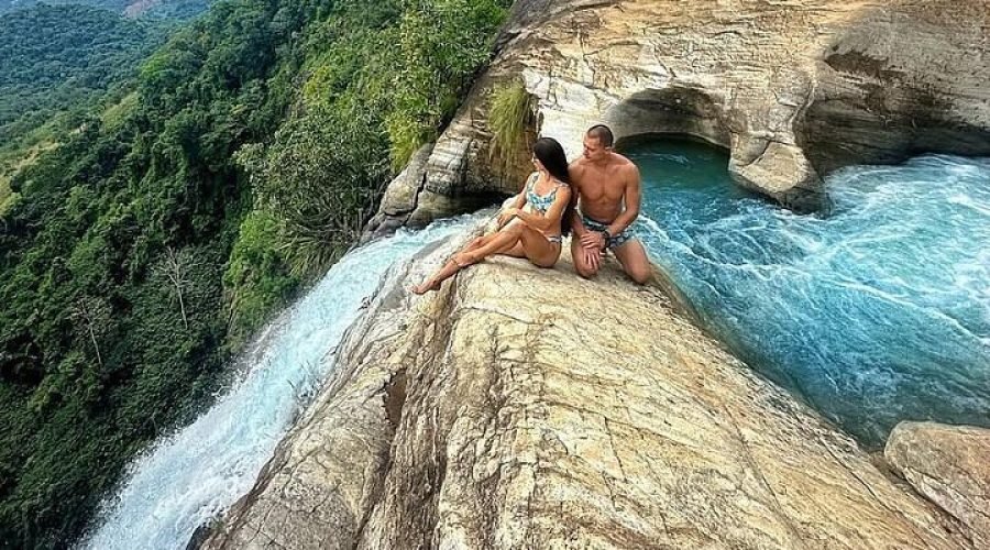 Couple sitting on a rocky ledge above a turquoise waterfall and lush forest valley.