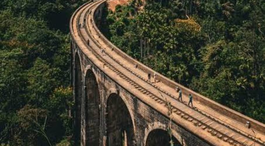 Hikers cross a curved stone railway viaduct surrounded by dense tropical forest.