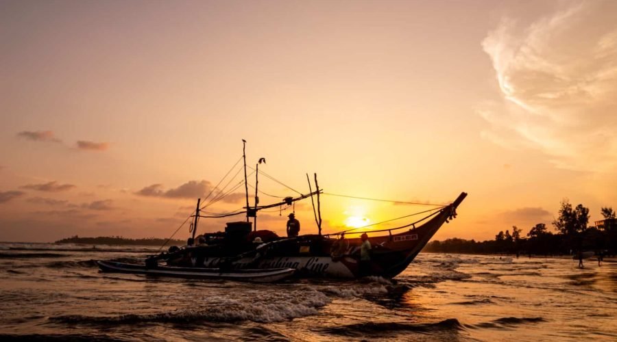 Silhouetted fishing boat glides across rippling water beneath a golden sunset sky.
