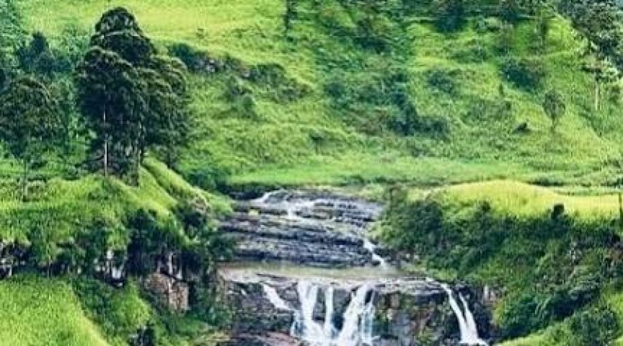Rocky waterfall cascading through lush green tea plantation hills in misty mountain scenery.