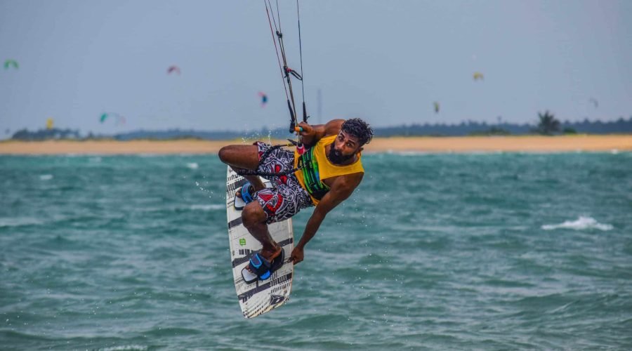 Kiteboarder performs an aerial trick above choppy turquoise water near a sandy shoreline.