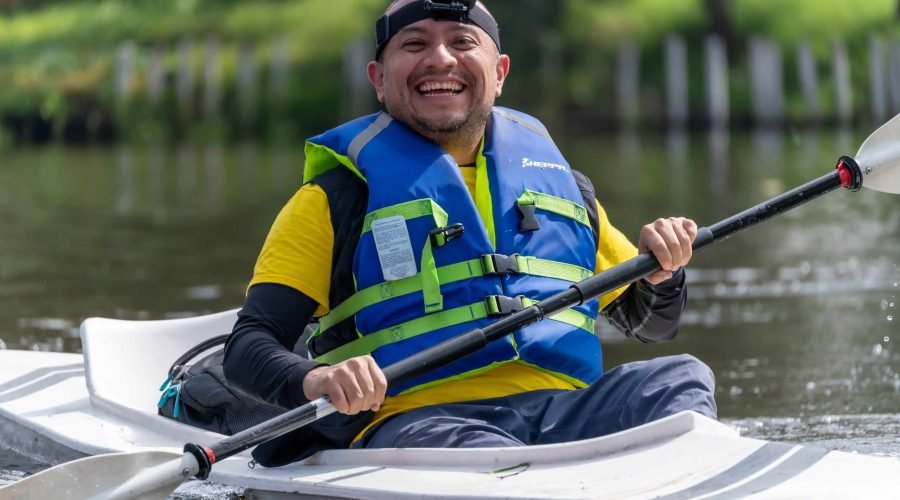 Smiling man paddles a white kayak across calm water wearing a blue life jacket.