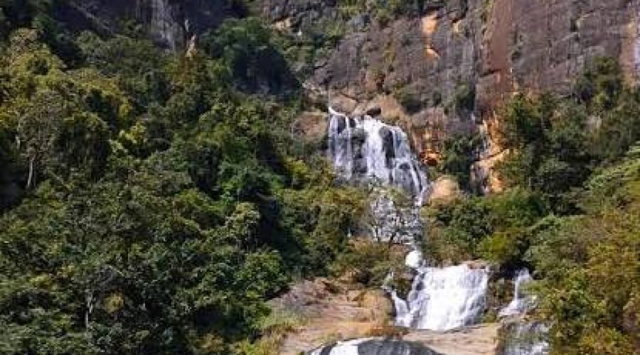 Multi-tiered mountain waterfall cascading through a lush forest gorge beneath rugged cliffs.
