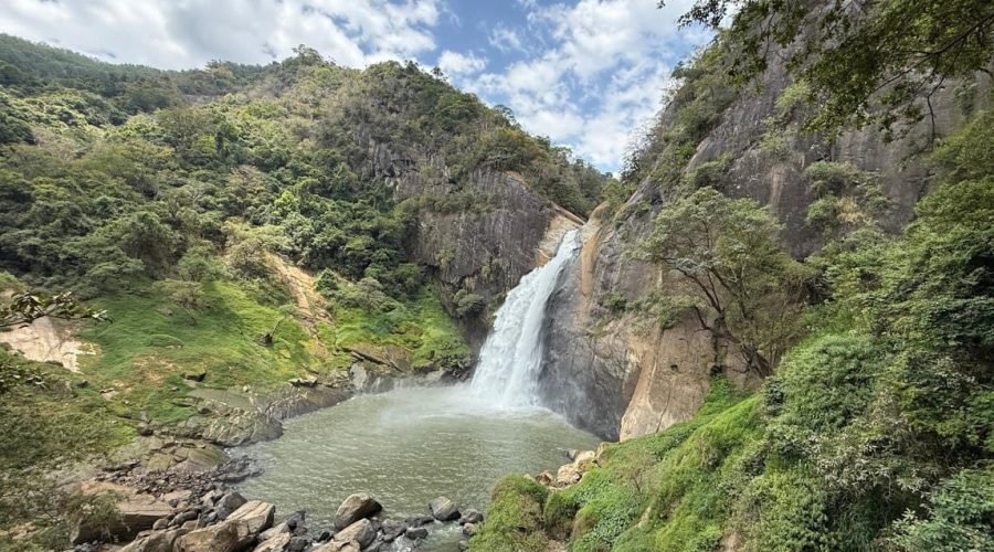 Mountain waterfall cascading into a pale green alpine pool surrounded by rugged cliffs and lush vegetation.