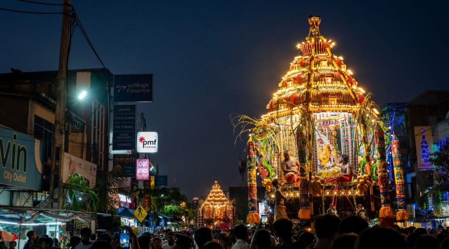 Illuminated religious chariot moves through a crowded street during a nighttime festival procession.