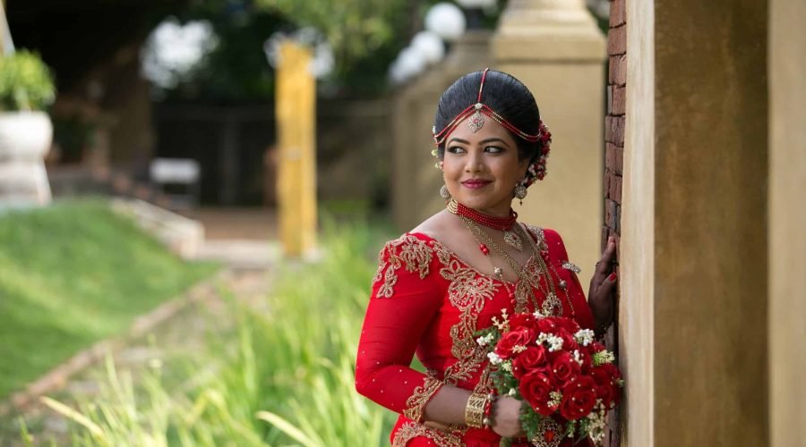 Bride in red traditional wedding attire holding a bouquet beside a stone column.