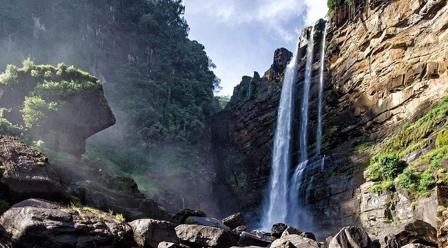 Majestic waterfall plunges down a rugged rocky cliff into a misty boulder-filled basin.