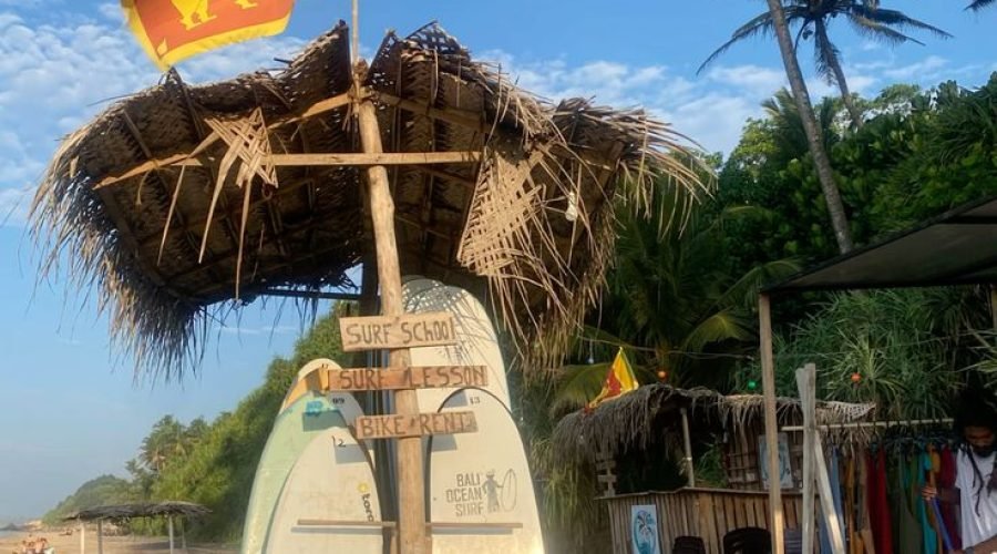 Sri Lanka surf beach with rustic signpost, surfboards, flag, palm trees, and beach shack.