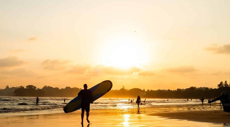 Lone surfer carrying a surfboard across a reflective beach at golden sunset.