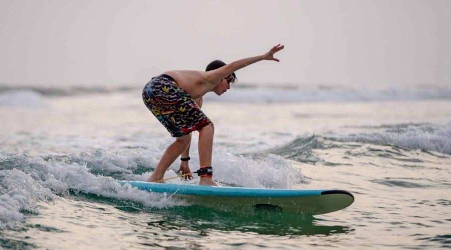Surfer crouches on a turquoise longboard while riding a small ocean wave.