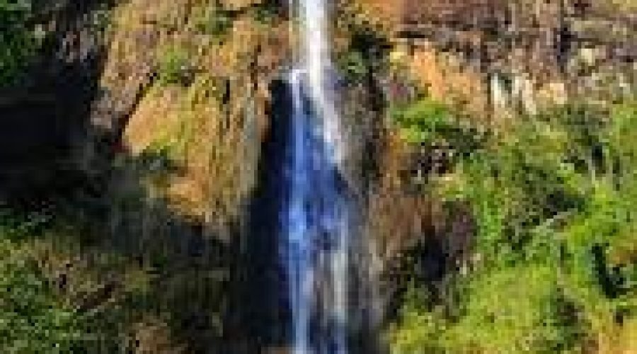 Tall tropical waterfall cascading down a sunlit rugged cliff surrounded by lush green vegetation.