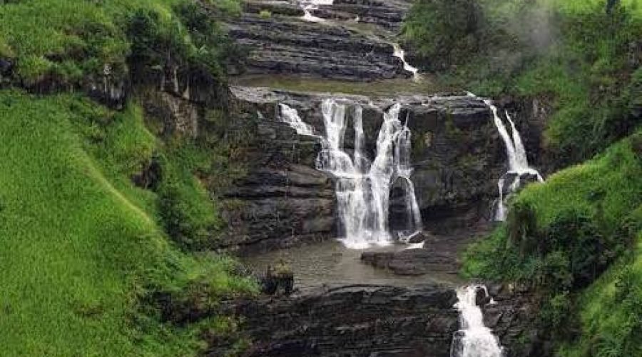 Tiered waterfall cascading over dark rocks through a lush, misty green hillside.