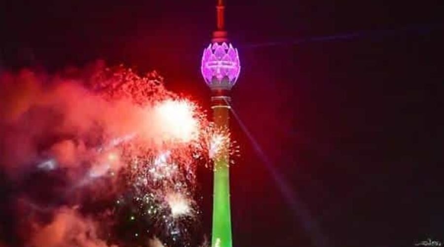 Green-lit observation tower beside colorful fireworks illuminating the night skyline.