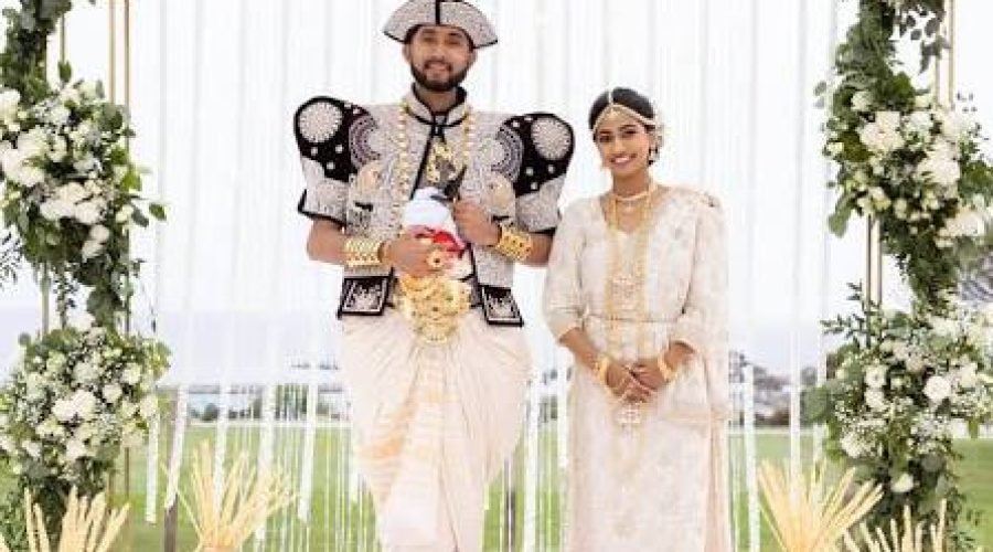 Newlywed couple in ornate traditional wedding attire beneath a white floral arch.