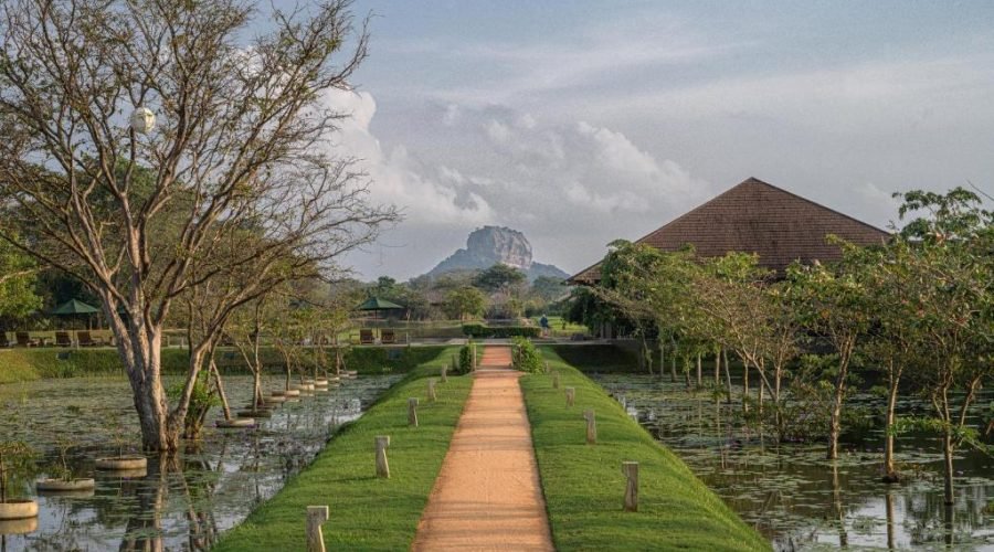 Tropical garden path crossing reflective waterways toward an ancient temple beneath a cloudy sky.