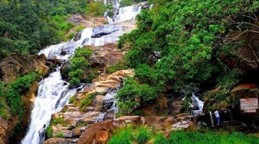 Multi-tiered tropical waterfall cascading through lush forest and rust-colored mountain rocks.