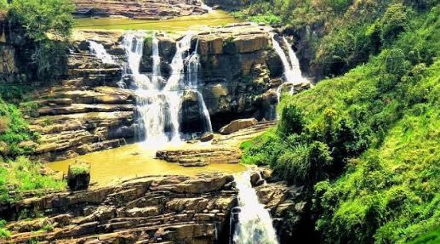 Multi-tiered tropical waterfall cascading over layered rocks amid lush green vegetation.