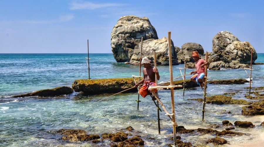 Tropical shoreline with turquoise water, rocky formations, wooden poles, and two people wading.