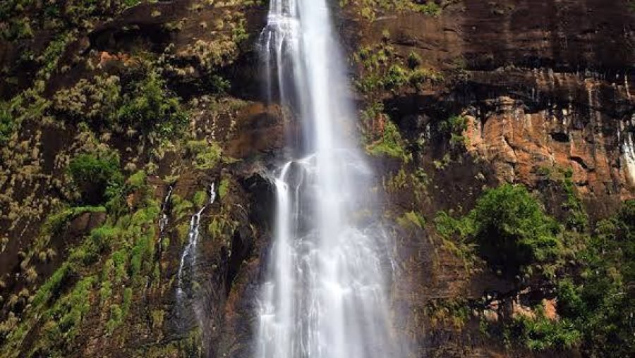 Tropical waterfall cascading down a rugged, vegetation-covered cliff under a deep blue sky.