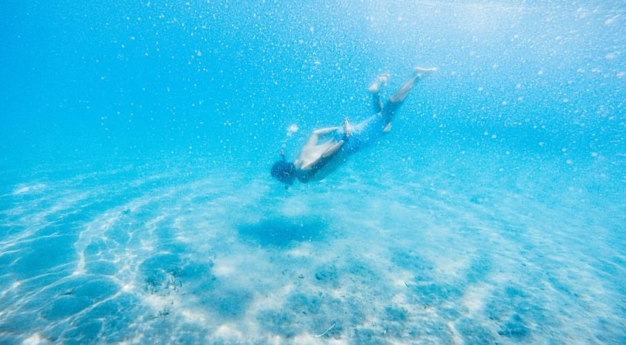 Snorkeler gliding above a sunlit sandy seabed in clear turquoise tropical water.