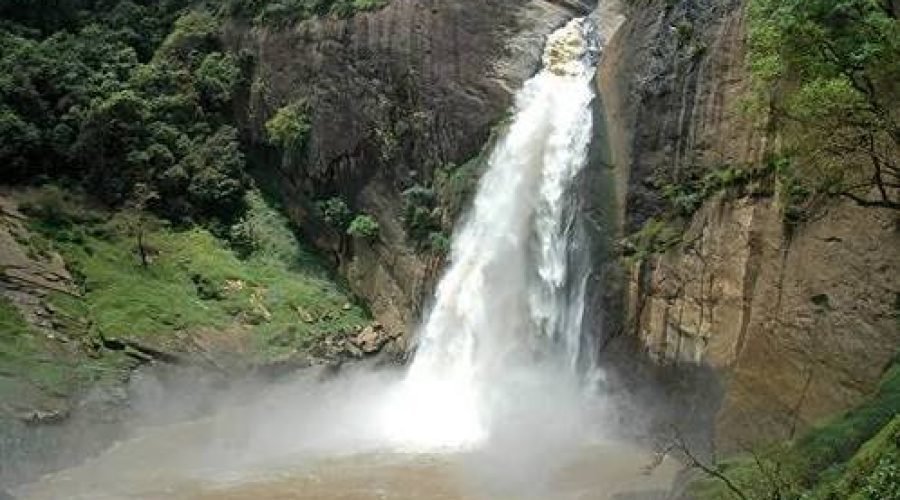 Powerful waterfall plunges through a lush rocky gorge into a misty brown river.