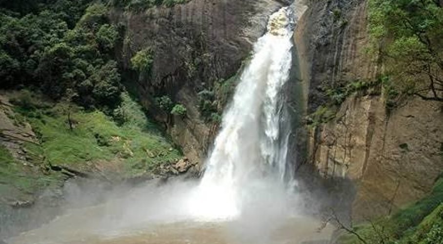 Powerful waterfall plunges through a lush rocky gorge into a misty brown river.