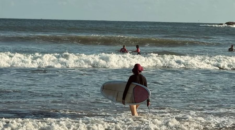 Surfer wades into ocean waves at Weligama Beach under a cloudy sky.