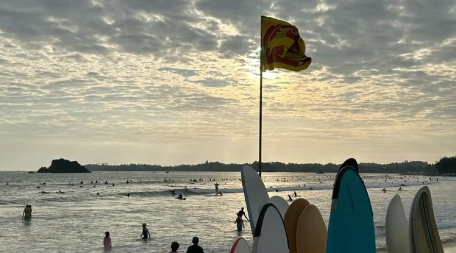 Surfboards beside a red-and-yellow warning flag on cloudy Weligama Beach, Sri Lanka.