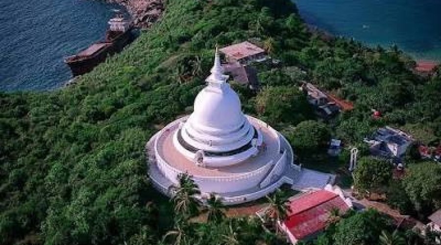 White Buddhist stupa rises above a lush tropical headland surrounded by blue ocean.